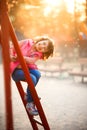 Little cute smiling 6 years old girl in jeans and pink vest sits on ladder in sunset time on playground in park. Child with active Royalty Free Stock Photo