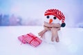 Little cute snowman in a red striped cap, a scarf with gift boxes on the snow on a winter day, close-up.