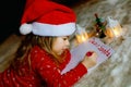 Little cute toddler girl in bed under Christmas tree, writing wish list letter to Santa Claus at home, indoors. Traditional