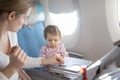 A little cute toddler girl sitting in an airplane in a chair by the porthole watching as mom wipes a folding table with