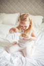 Little cute white-haired girl eats granola on the bed in her bright room. Healthy food. Home comfort Royalty Free Stock Photo