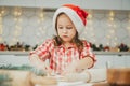 Dark-haired girl 3 years old in red Christmas cap and checkered shirt cuts out gingerbread cookies from rolled dough Royalty Free Stock Photo