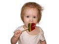 A little disheveled blue-eyed girl baby in white body tastes candy on a stick, a lollipop Royalty Free Stock Photo