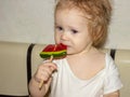 A little disheveled blue-eyed girl baby in white body tastes candy on a stick, a lollipop Royalty Free Stock Photo