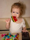 A little disheveled blue-eyed girl baby in white body tastes candy on a stick, a lollipop Royalty Free Stock Photo