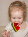A little disheveled blue-eyed girl baby in white body tastes candy on a stick, a lollipop Royalty Free Stock Photo