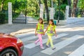 Little elementary school kids crossing the street wearing a vest with the stop sine on it.