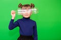 Little environmentalist girl isolated on green background looking through plastic bottle and shows interest in reducing plastic