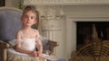 The little fair-haired girl sits on a chair at a fireplace in a white tutu. Darlings small in white packs.