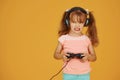 Little female gamer in headphones and with joystick in hands playing video games against yellow background