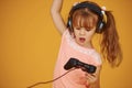 Little female gamer in headphones and with joystick in hands playing video games against yellow background