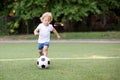 Little football player: blonde child in white shirt and blue shorts running along the green soccer field ready to kick ball. Royalty Free Stock Photo