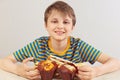 Little funny boy in a striped shirt at the table with muffins and sandwich on white background
