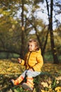 Little funny girl in stylish yellow warm jacket, jeans, orange rubber boots sits on old tree stump in autumn forest or park Royalty Free Stock Photo