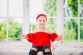 Little funny two years old girl child in stylish red clothes and jeans and sneakers on windowsill near window. With a smile On his Royalty Free Stock Photo