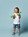 Ginger baby boy or girl in white t-shirt, shoes, denim shorts. Smiling, holding two green dumbbells, posing on blue background Royalty Free Stock Photo