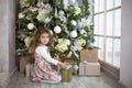 A little girl in a beautiful dress is sitting under a Christmas tree with a gift box and a bow. The light from the large window, t Royalty Free Stock Photo