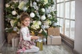 A little girl in a beautiful dress is sitting under a Christmas tree with a gift box and a bow. The light from the large window, t Royalty Free Stock Photo