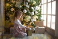 A little girl in a beautiful dress is sitting under a Christmas tree with a gift box and a bow. The light from the large window, t Royalty Free Stock Photo