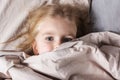 Little girl with beautiful eyes lying in bed. The baby is hiding under a blanket.