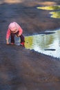 Little girl bending to the ground playing in big spring puddle wearing purple nylon and pink bucket hat