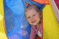 A little girl with blond curly hair looks out from the multicolored tent and smiles