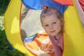 A little girl with blond curly hair looks out from the multicolored tent and smiles