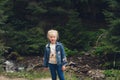 A little girl with blond hair, in jeans clothes, stands against the backdrop of a mountain river Royalty Free Stock Photo