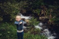 A little girl with blond hair, in jeans clothes, stands against the backdrop of a mountain river with stones Royalty Free Stock Photo