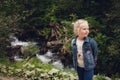 A little girl with blond hair, in jeans clothes, stands against the backdrop of a mountain river with stones Royalty Free Stock Photo