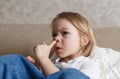 a little girl in a blue denim jumpsuit sits on the couch and picks her nose. Close-up.