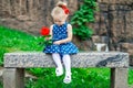 Little girl in a blue dress with white polka dots sits on a bench with a rose