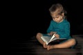 a little girl in a blue dress 4 years old is sitting on the floor in a dark room and reading a book. home schooling in quarantine Royalty Free Stock Photo