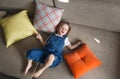 a little girl in a blue jumpsuit is lying at home on the sofa among the colorful pillows on her back.