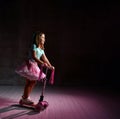 Little girl in blue t-shirt, poofy skirt and white sneakers. She is smiling and riding a scooter on black background. Full length