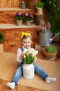 Little girl with a bouquet of white tulips sits on porch of a wooden house, around green houseplants and flowers. Childhood concep