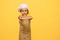 little girl in chef hat and apron, smiles and holds out at camera a wooden board, isolated over yellow studio background Royalty Free Stock Photo