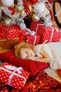 A little girl with curly blonde hair in a white dress lies on the floor on a pillow near the gifts and looks at the camera against