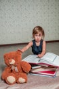 Little girl in dark blue dress reading book sitting on the floor near teddy bear. Child reads story for toy. Royalty Free Stock Photo