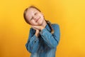 Little girl in a denim shirt on a yellow background. The child closed her eyes and laid her head on folded palms