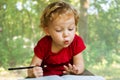 Little girl draws in a notebook on a background of nature. The beautiful, emotional face of a child of three years