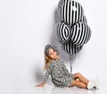 Little girl in dress, headband and sneakers. She smiling, holding striped balloons, sitting on floor, isolated on white. Close up Royalty Free Stock Photo