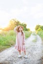 Little girl in dress and straw hat is walking along country road. Happy childhood. Summer vacation. Kid walks along rural road on Royalty Free Stock Photo