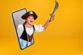 A little girl dressed as a pirate for Halloween holds a saber and looks from a smartphone. Child online on a mobile phone screen Royalty Free Stock Photo