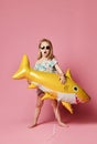 Girl wears a balloon in the shape of a yellow shark-fish, celebrates a holiday, smiles broadly, stands against a pink background