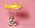 Little girl, dressed in a summer dress, wears a balloon in the shape of a yellow shark-fish, stands against a pink background