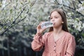 A little girl drinks clean fresh water from a plastic bottle near a flowering tree in the garden