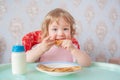 Little girl eating pancakes with milk in a pink bib. Cute little baby eating pancakes. The baby eats independently. Sitting in a Royalty Free Stock Photo