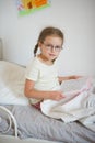 Little girl with glasses sitting in bed and holding an open book.