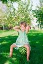 Little girl in green dress barefoot sitting on watermelon in the park.